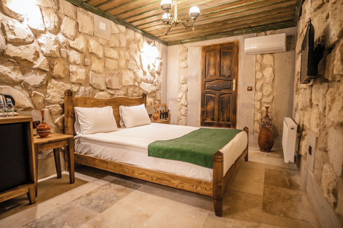 A stone-walled bedroom with a wooden bed, a bedside table, and a small table with a vase.