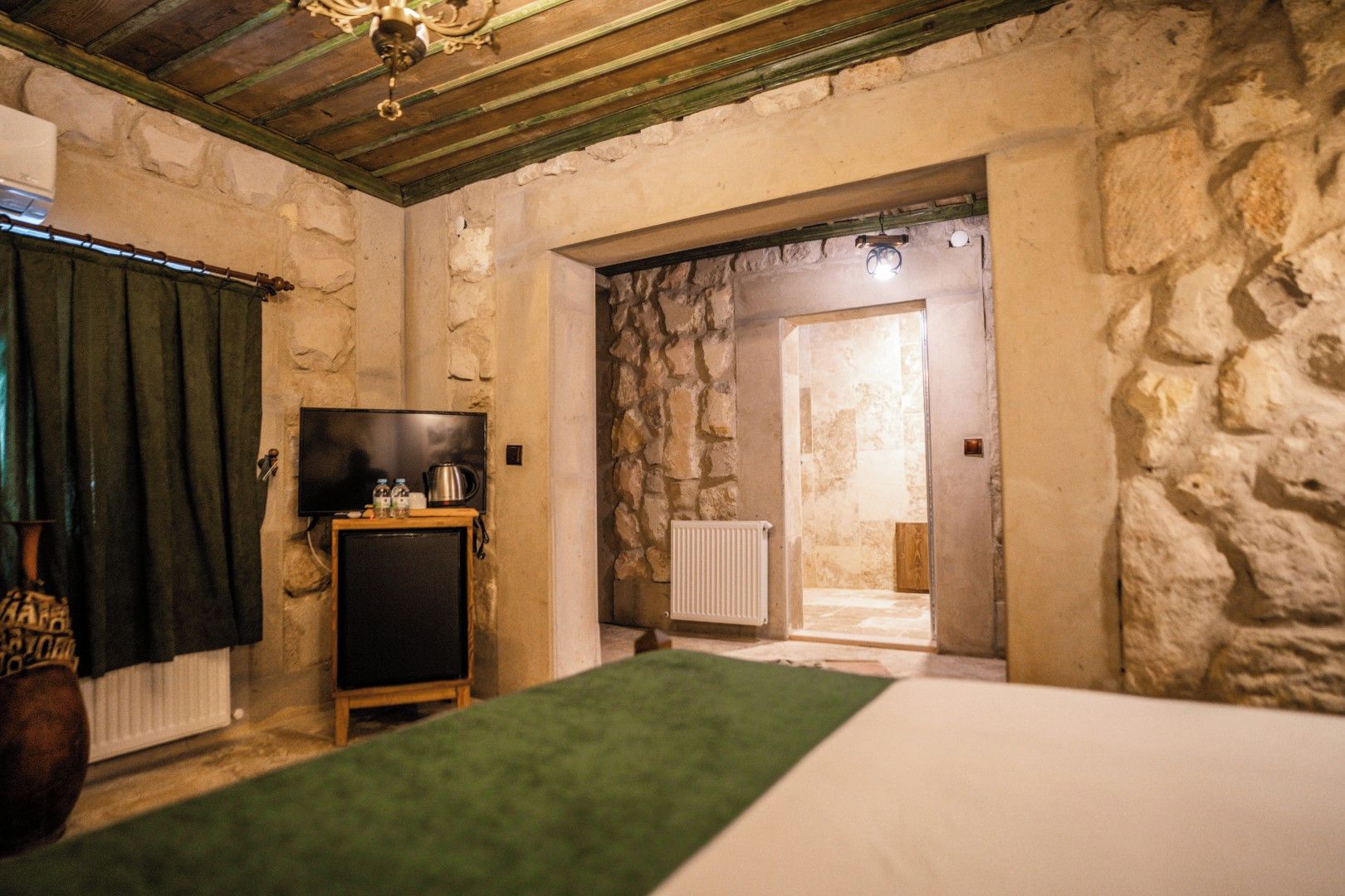 Una habitación con pared de piedra, un televisor sobre un mueble de madera, un radiador y un acceso a otra habitación.