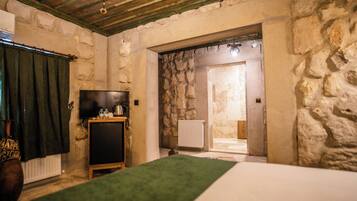 Una habitación con pared de piedra, un televisor sobre un mueble de madera, un radiador y un acceso a otra habitación.