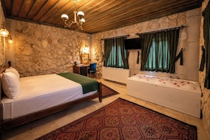 A bedroom with a bed, a desk, a chair, a bathtub with rose petals, and a stone wall. - Vista Rosa Cappadocia (Çavuşin)