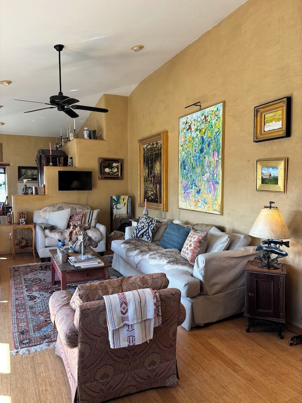 A cozy living room with a sofa, armchairs, a coffee table, and a fireplace. There are framed pictures on the wall and a ceiling fan.