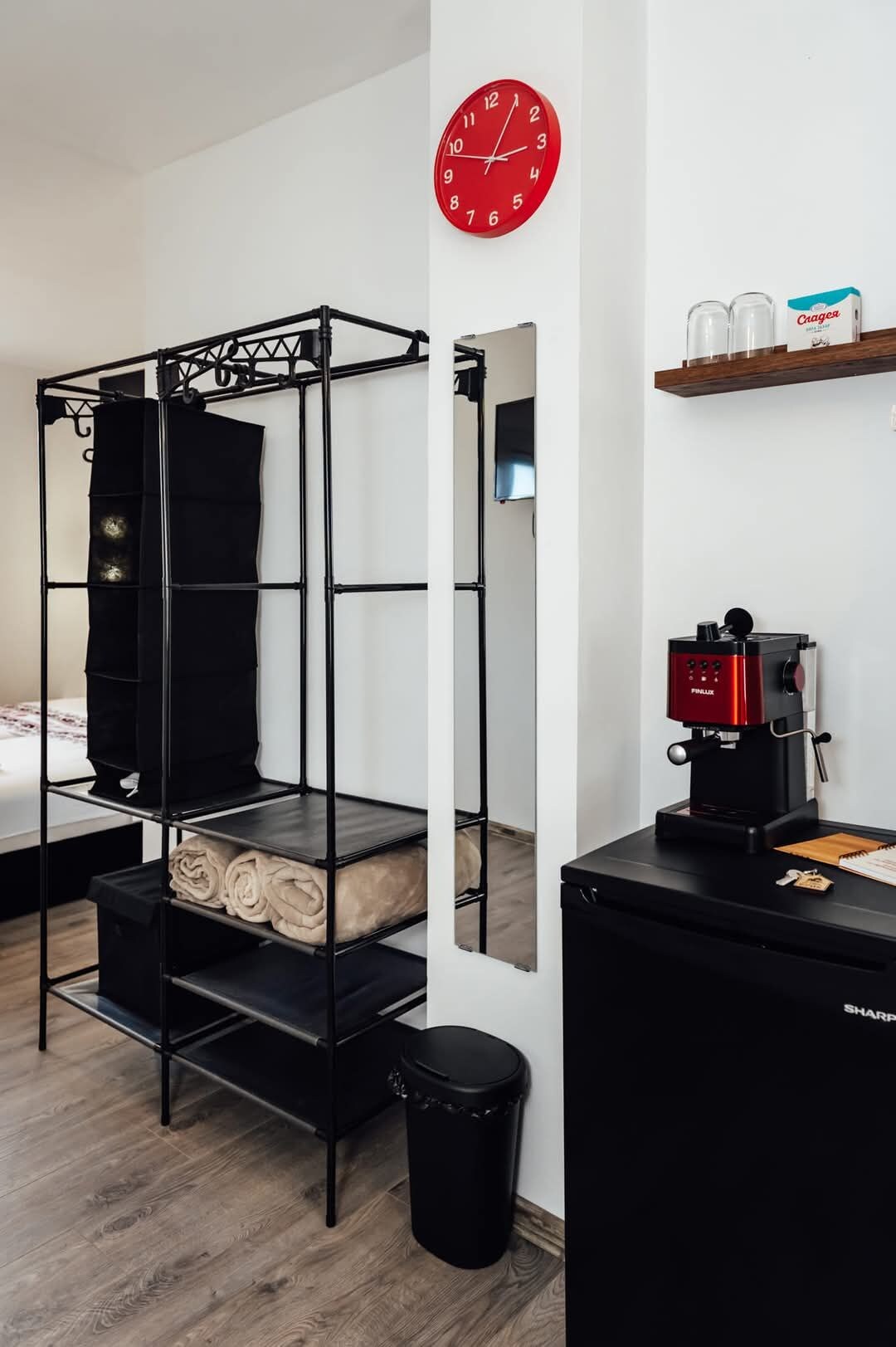 A room with a bunk bed, a red wall clock, a coffee machine, and a trash bin.