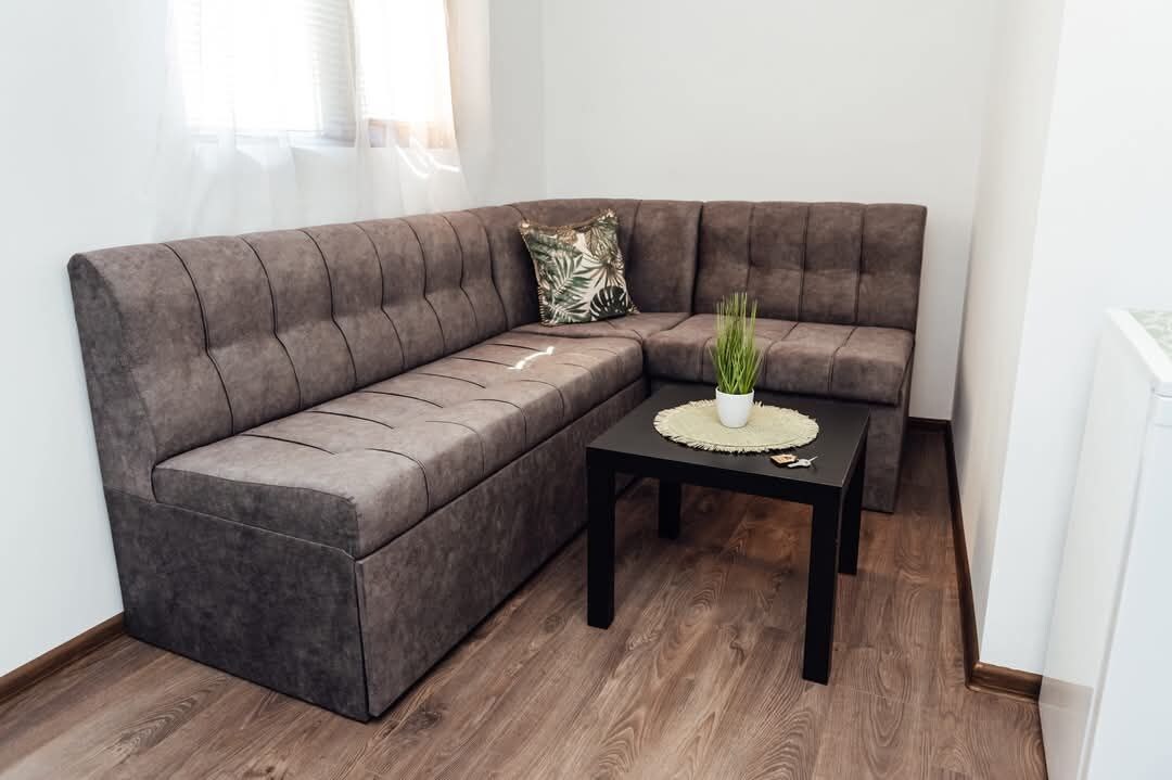 Ein Eckwohnzimmer mit einer grauen Eckcouch, einem kleinen schwarzen Couchtisch mit einer Pflanze und einem gemusterten Kissen.