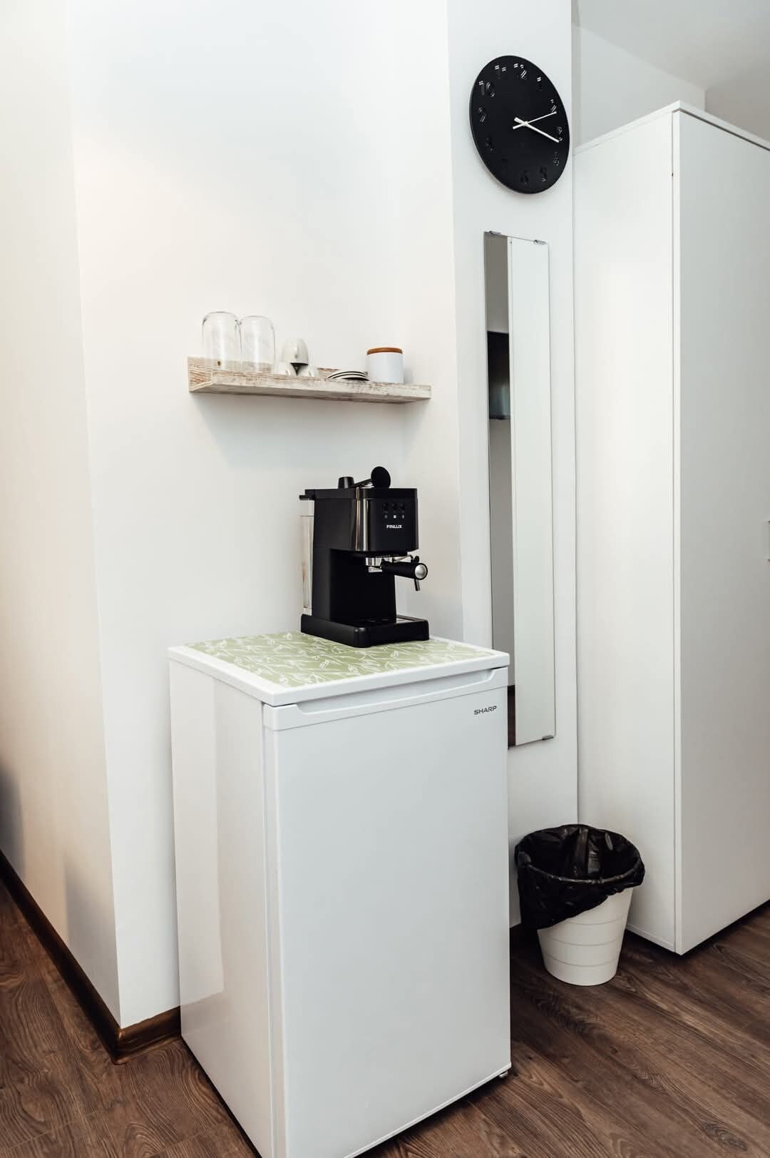 Eine kleine Küchenzeile mit Kaffeemaschine, kleinem Kühlschrank, Mülleimer, einer Ablage mit Tassen und Behälter sowie einer Wanduhr.