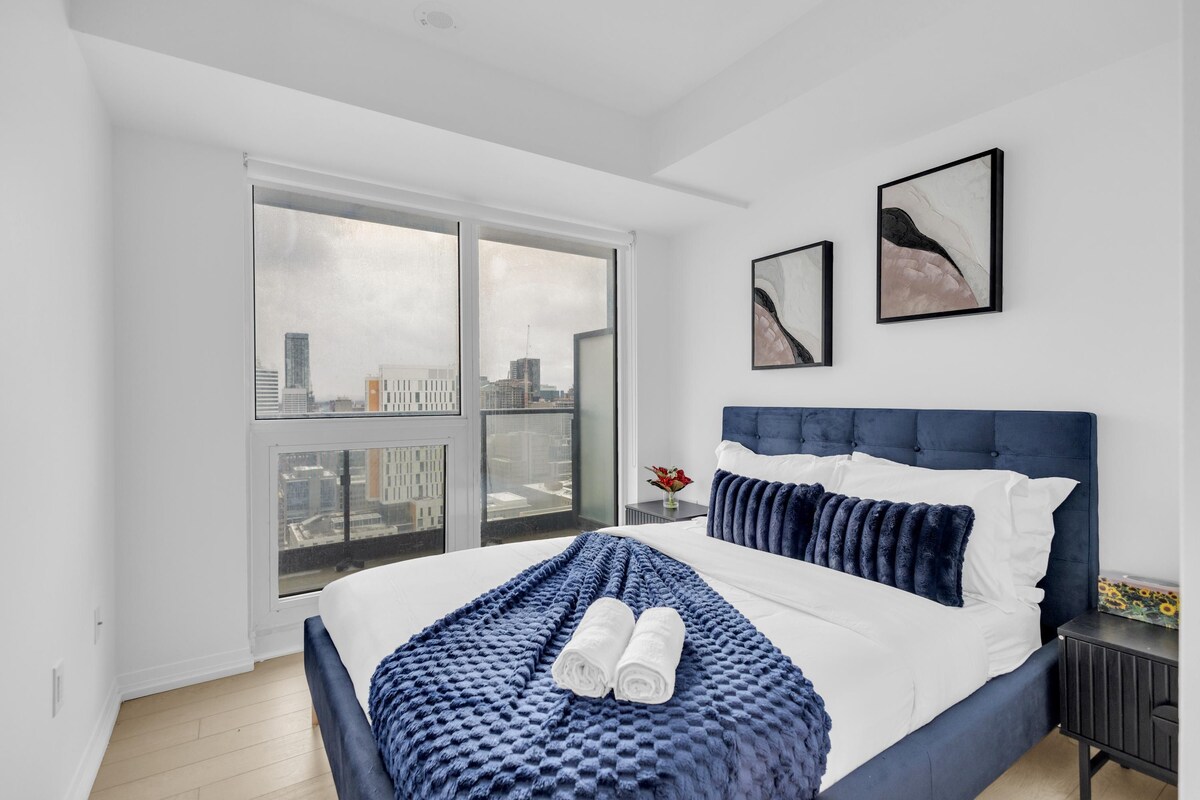 Een moderne slaapkamer met een groot bed, uitzicht op de stad door middel van raam-tot-plafond ramen en twee ingelijste kunstwerken aan de muur.
