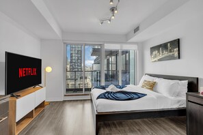 Une chambre moderne avec un grand lit, un téléviseur affichant le logo de Netflix, une vue sur la ville par la fenêtre et un tableau au mur.
