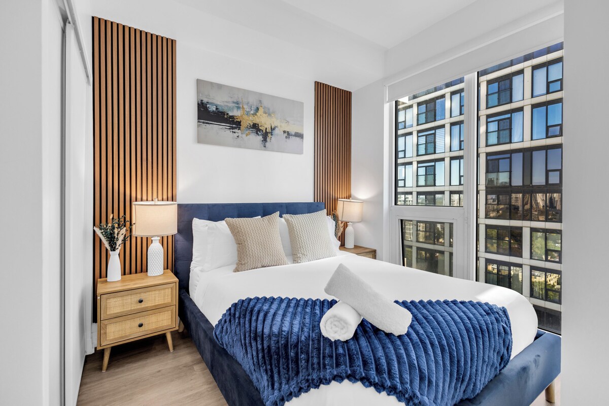 Een moderne hotelkamer met een groot bed, houten lambrisering, een stadsgezicht schilderij en uitzicht op een gebouw door het raam.
