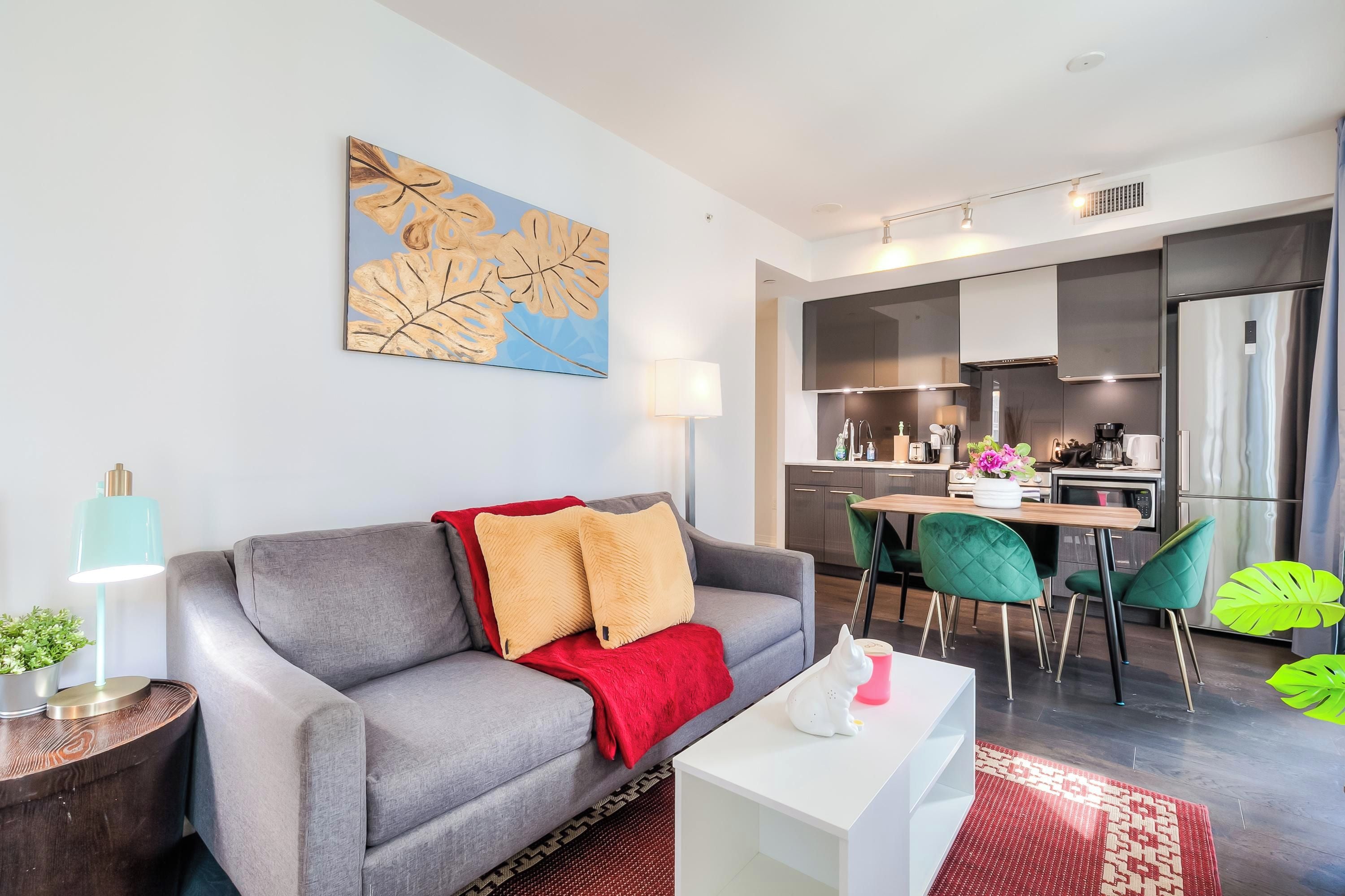 Una sala de estar moderna con un sofá gris, una manta roja y una mesa de centro blanca. Hay una zona de cocina con una mesa de comedor y sillas, y un amplio ventanal que proporciona luz natural.
