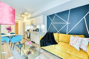 A modern living room with a yellow sofa, a glass coffee table, and a dining area with blue chairs. - Dundas Suites - Heart of Downtown (Toronto)