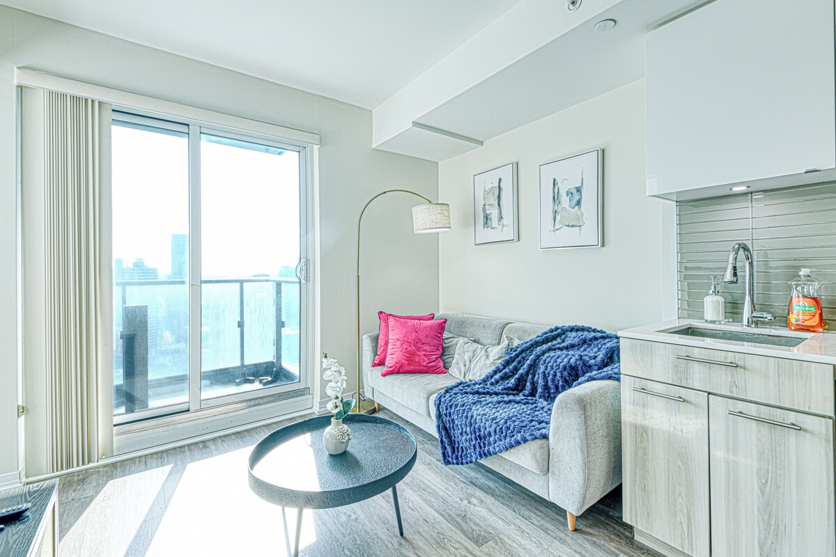 Een moderne woonkamer met een groot raam, een bank met een plaid, een kleine ronde tafel met een vaas, en een keukenhoek met een spoelbak en kastjes.