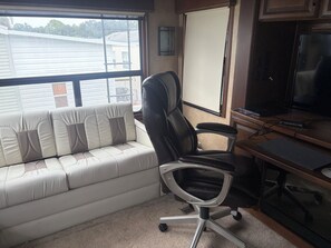 Quarto com sofá branco, cadeira de escritório preta, televisão e escrivaninha com computador.