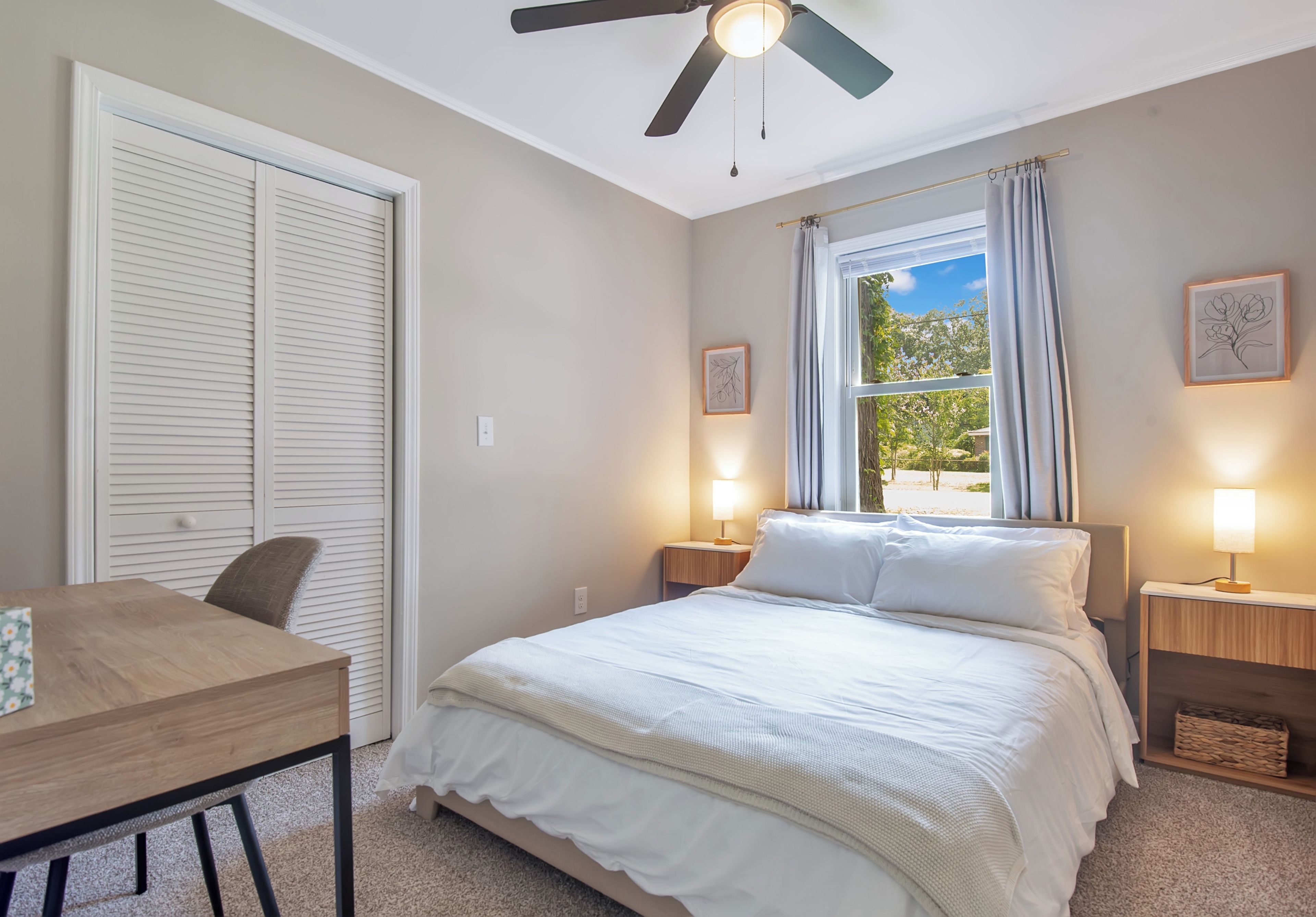 A bedroom with a bed, bedside lamps, a ceiling fan, a window with curtains, and a desk with a chair.