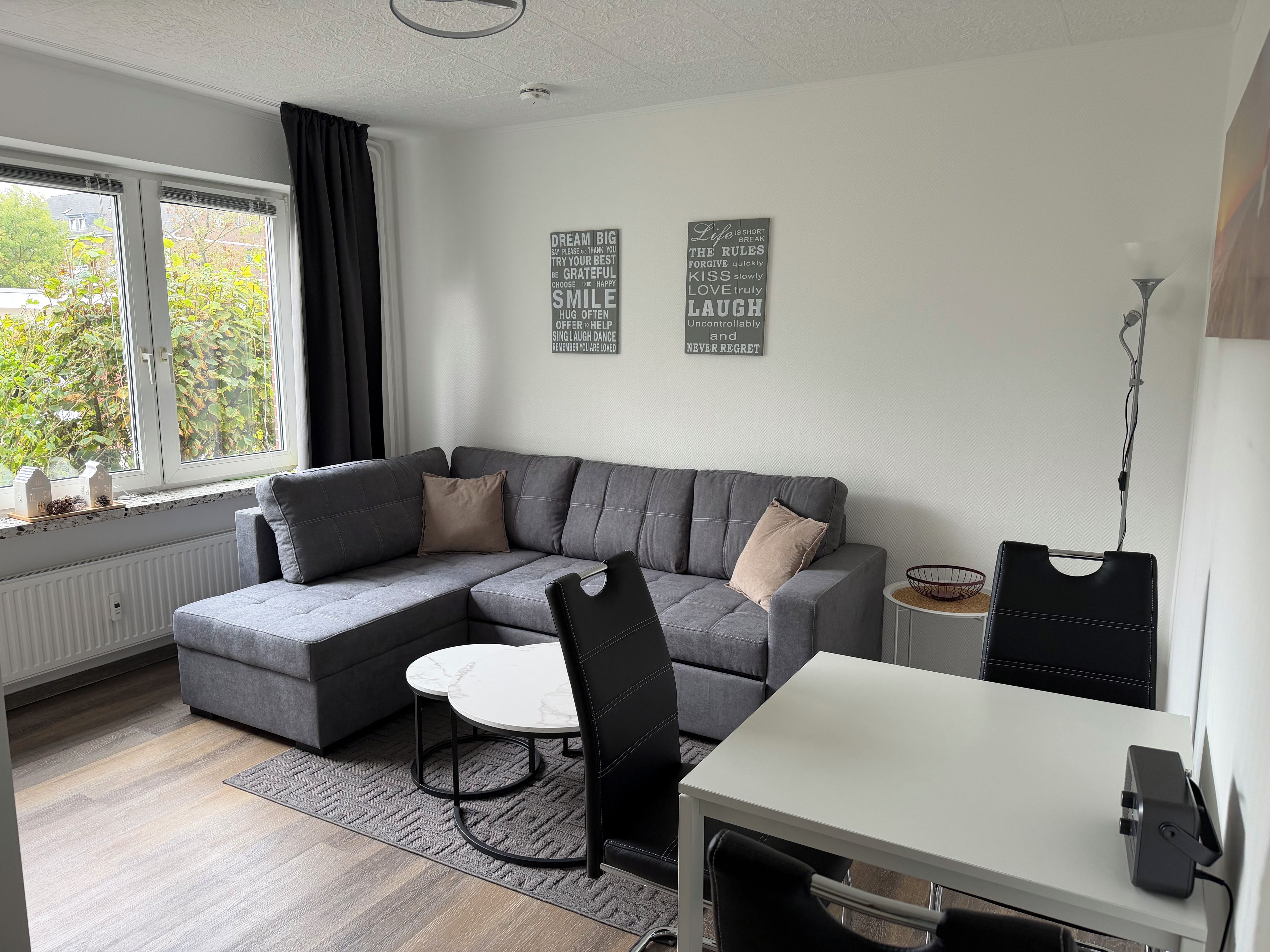A modern living room with a grey sectional sofa, a small round table, and a black chair. There are two wall decorations with inspirational quotes.