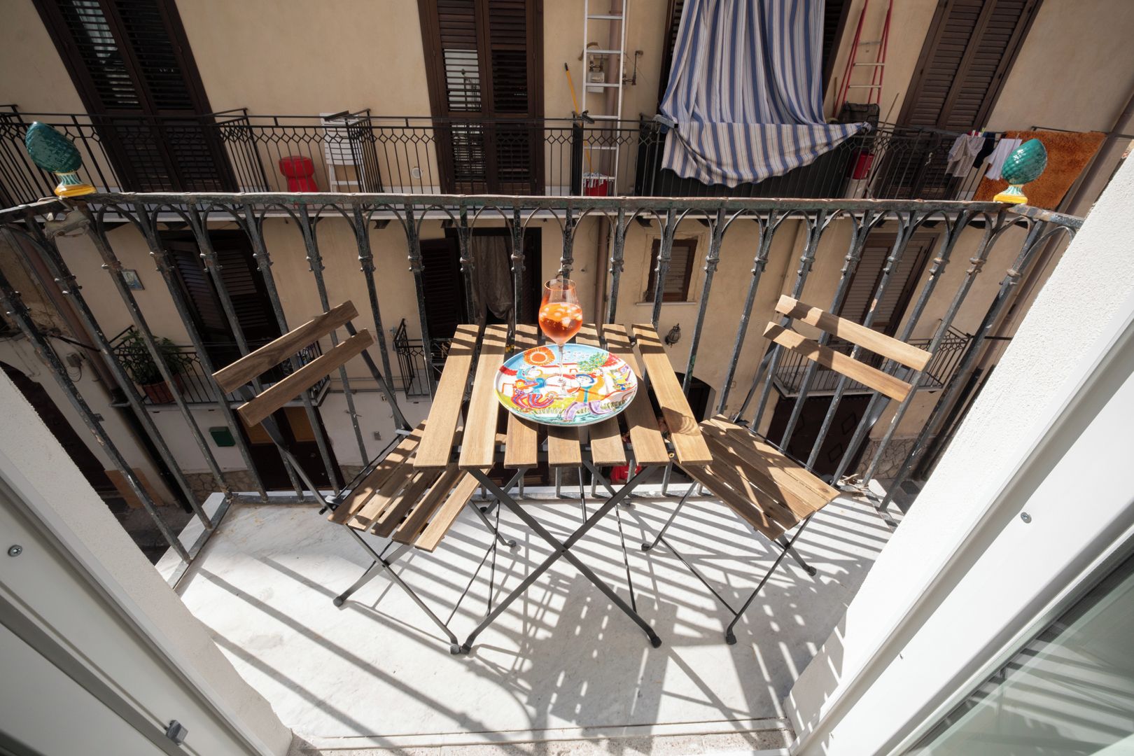 Un balcon avec une table et des chaises en bois, une assiette colorée et un verre de jus d’orange.