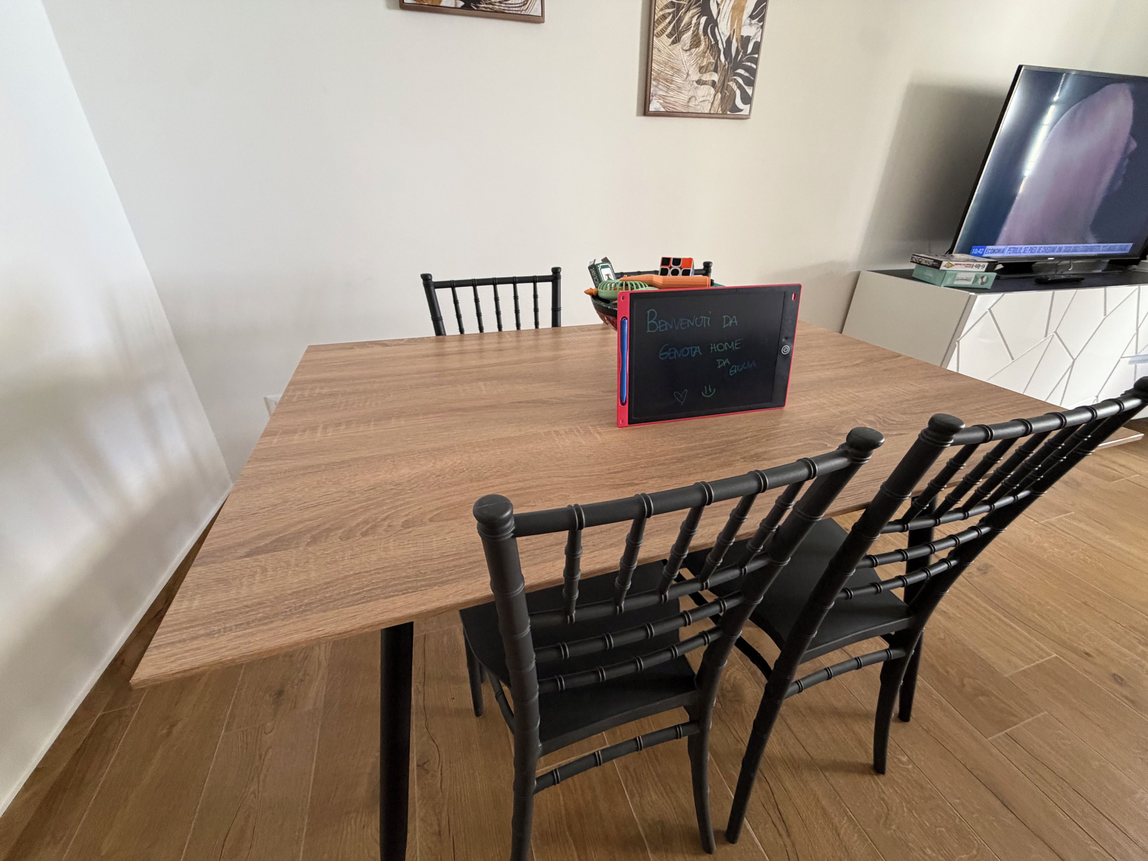 Un espace repas avec une table en bois, des chaises noires, un tableau noir sur lequel est écrit un message de bienvenue et un téléviseur posé sur un meuble.