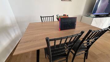 Un espace repas avec une table en bois, des chaises noires, un tableau noir sur lequel est écrit un message de bienvenue et un téléviseur posé sur un meuble.