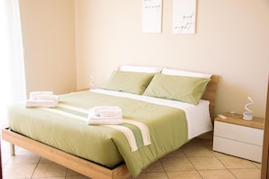 Cama bem arrumada com roupa de cama verde e almofadas brancas, mesa de cabeceira com abajur e duas obras de arte na parede com texto.
