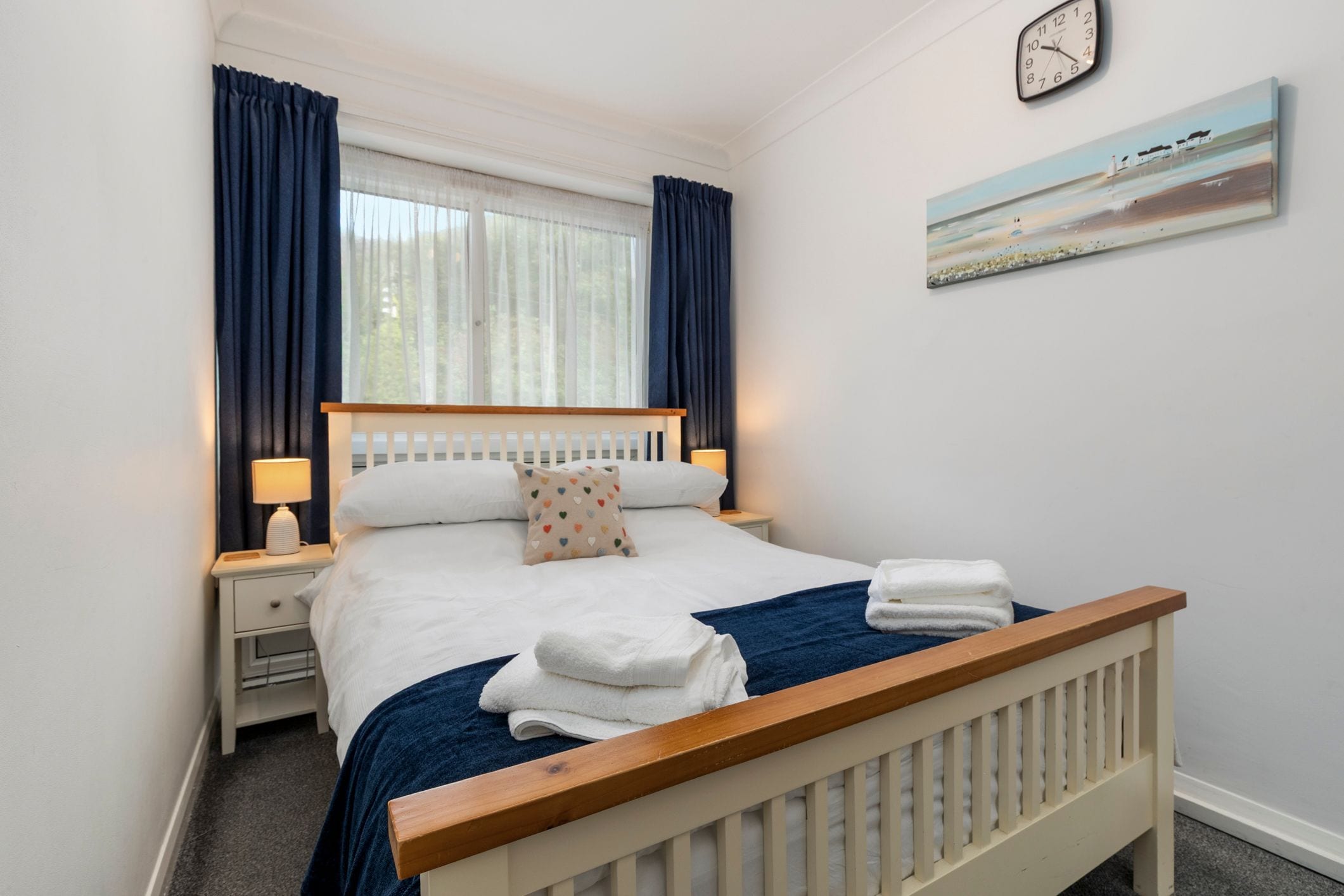 A neatly made bed with white linens and a blue blanket, two bedside tables with lamps, a clock on the wall, and a painting above the bed.