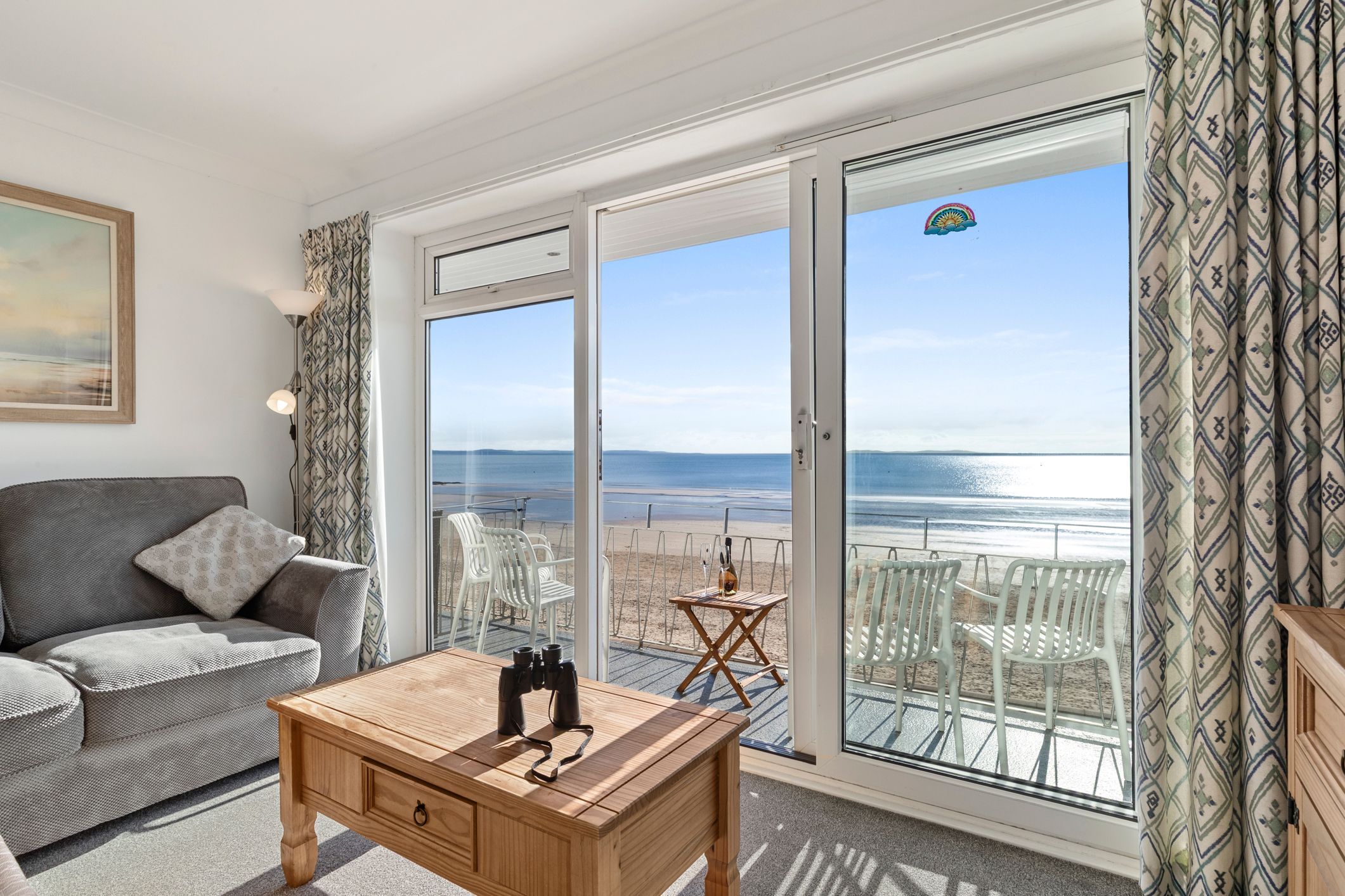 A room with a sofa, a wooden table with binoculars, and a view of the beach through large glass doors.
