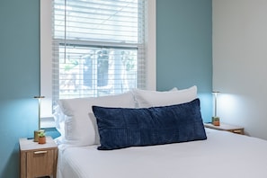 Un dormitorio con una cama, almohadas blancas, una almohada azul oscuro, una mesita de noche con una lámpara y una ventana con persianas.