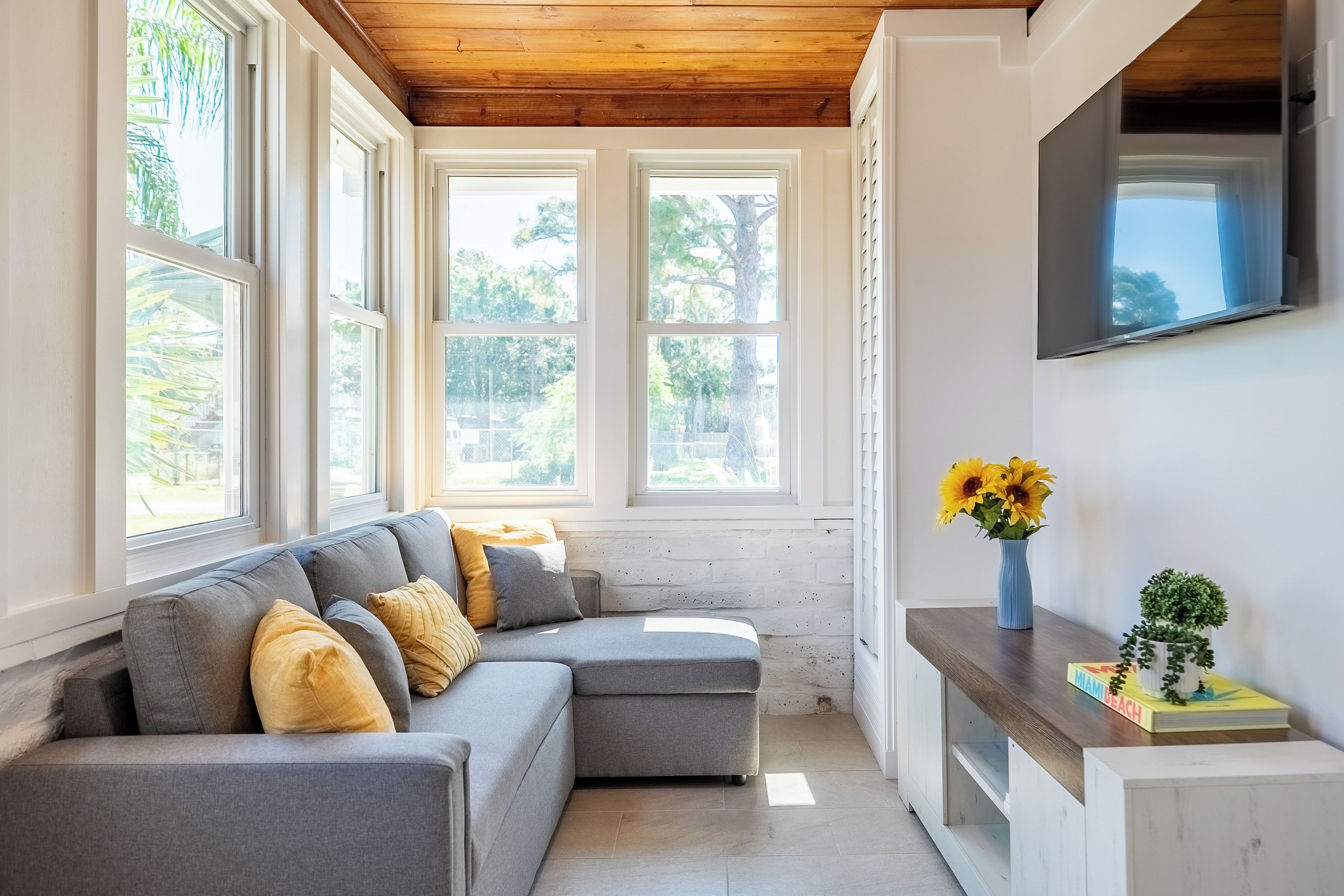 A room with a grey sectional sofa, a wooden ceiling, and a TV mounted on the wall.