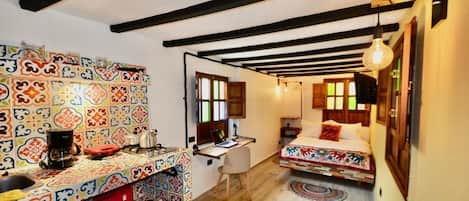 A room with a colorful tiled backsplash, a bed with a patterned bedspread, a desk with a computer, and a red Coca-Cola refrigerator.