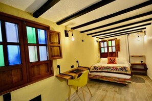 Un dormitorio con cama, escritorio y silla. Hay ventanas con vitrales, un techo con vigas a la vista y una mesita de noche con lámpara.