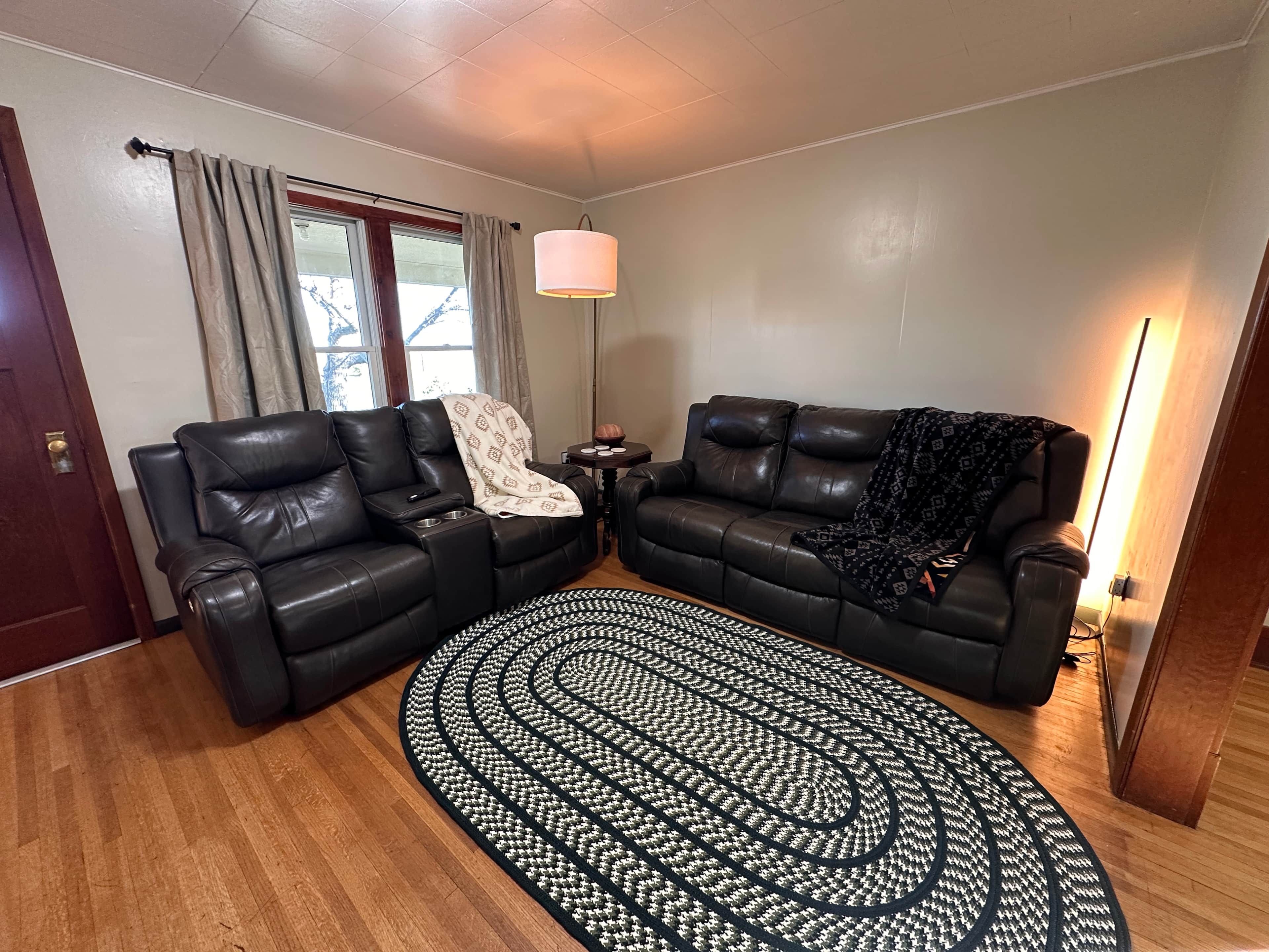A living room with a black leather sofa, a patterned rug, a lamp, and a window with curtains.