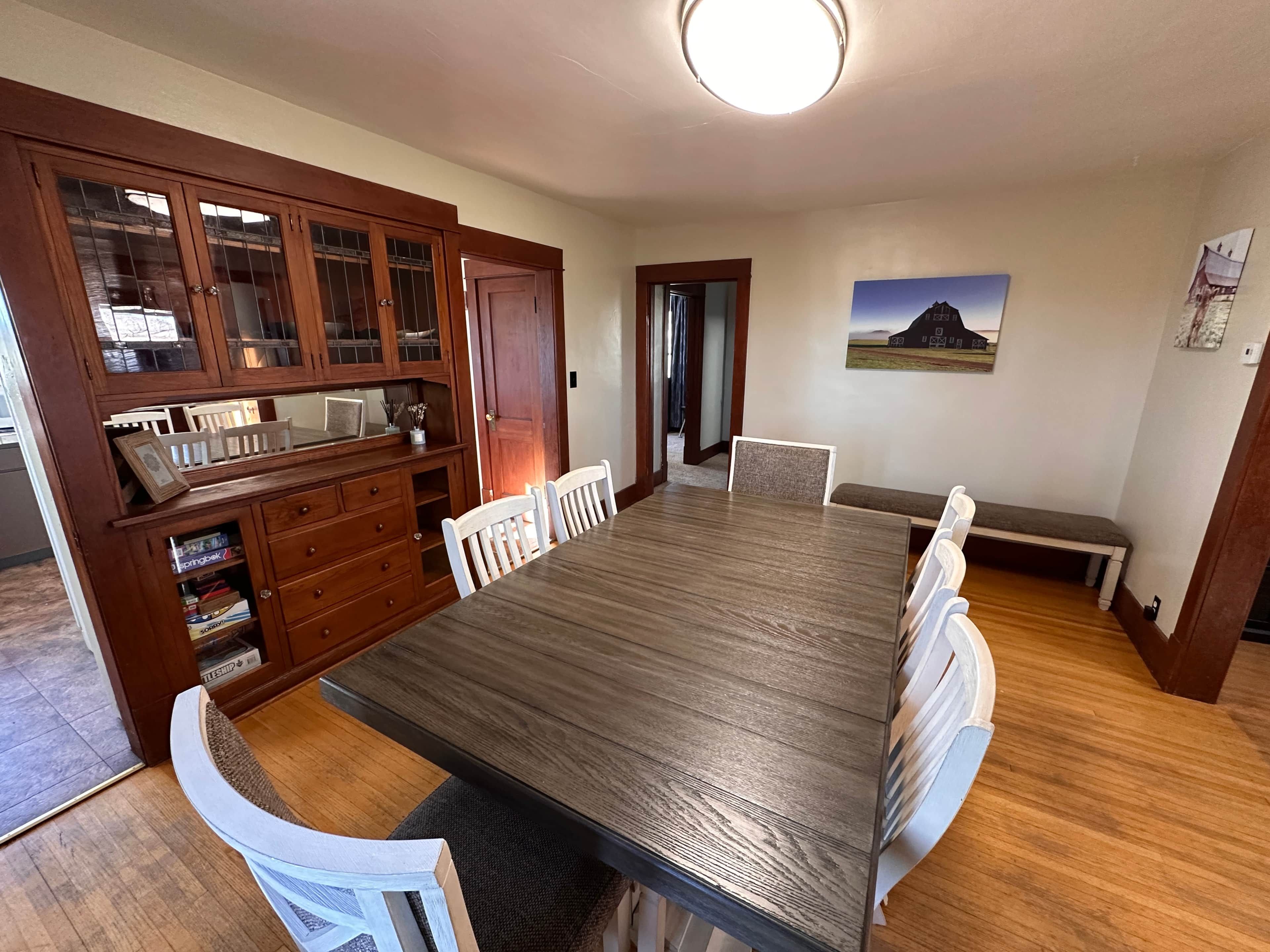 Une salle à manger avec une grande table en bois entourée de chaises, un buffet en bois intégré avec des portes en verre, et un banc le long d’un des murs.