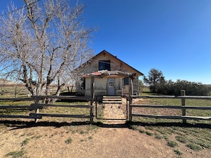 A rustic wooden cabin with a red roof, surrounded by a wooden fence and a tree with bare branches.