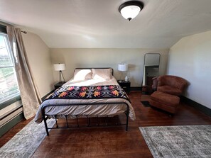 Ein Schlafzimmer mit Bett, Nachttischlampen, einem Sessel, einem Spiegel und einem Fenster mit Vorhängen.