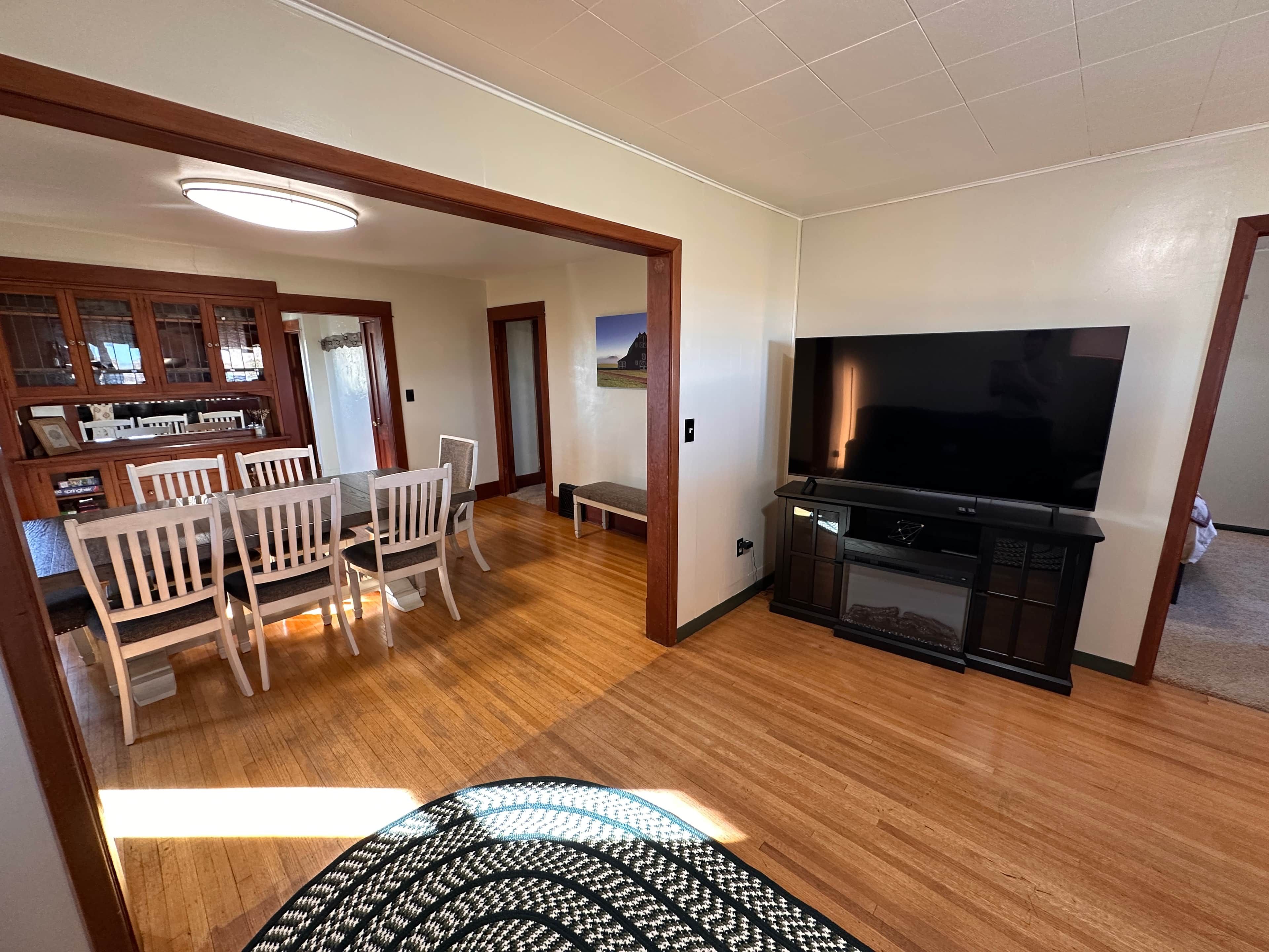 A living room with a wooden floor, a dining area with white chairs, a TV stand, and a kitchen area with wooden cabinets.