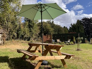 Ein Picknicktisch mit Vordach, umgeben von einer grünen Wiese mit Bäumen und einem Gebäude im Hintergrund.