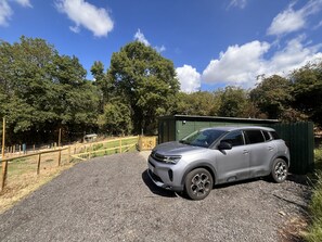En silverfärgad SUV står parkerad på en grusväg, med ett grönt förråd i bakgrunden, omgiven av träd och ett trästaket.