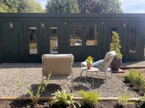 Um edifício verde com várias janelas e portas, uma cadeira bege, uma planta em vaso e uma pequena área de jardim com pedriscos e plantas.