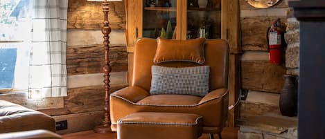 A log cabin interior with a leather armchair, a matching ottoman, a wooden floor, a fireplace, and a glass-fronted cabinet with decorative items.