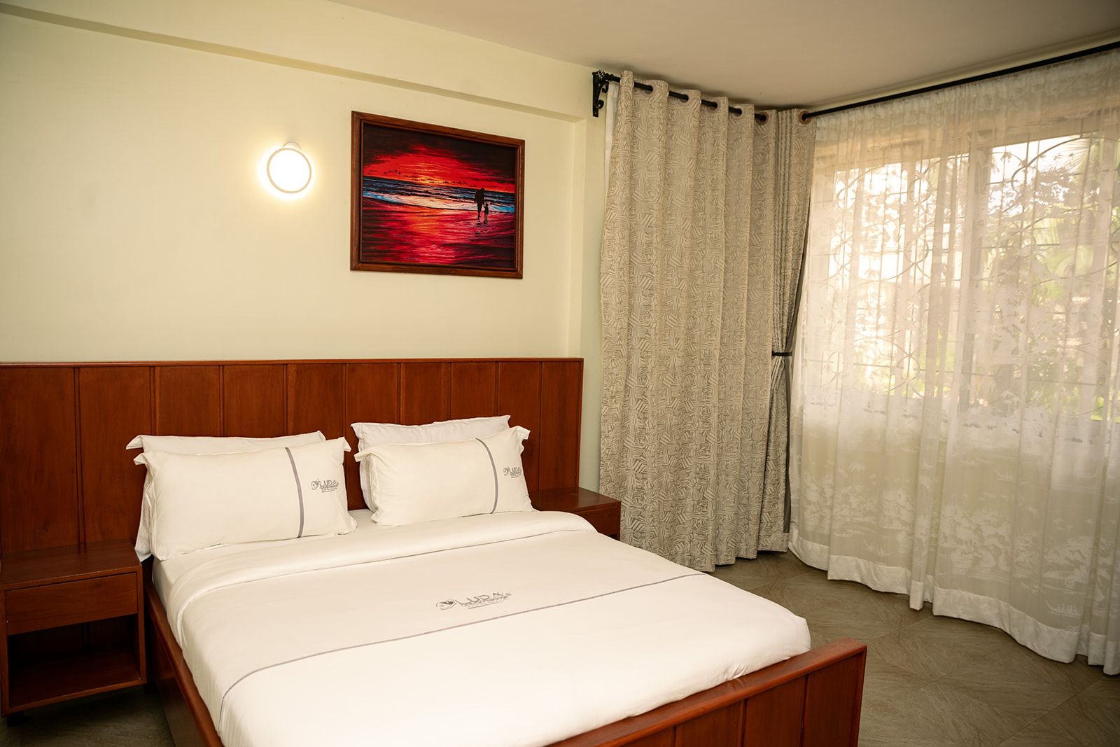 A hotel room with a bed, a nightstand, a painting, and a window with curtains.