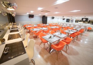 A dining area with multiple tables and chairs, a kitchen area in the background, and a modern interior design. - Goa Essence Citymix (Campina Grande)