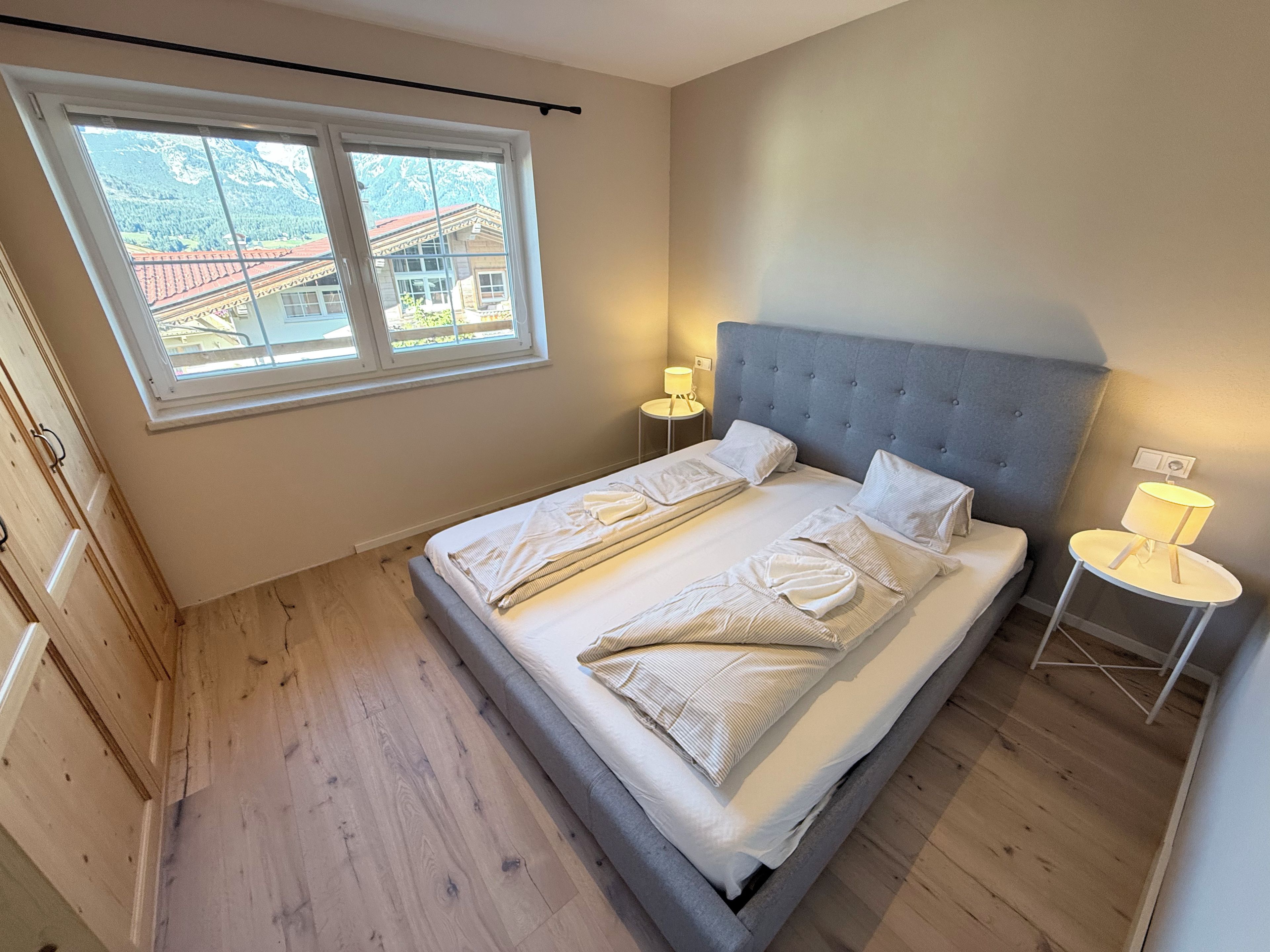 A bedroom with a large bed, two bedside tables, a window with a view of mountains, and wooden flooring.