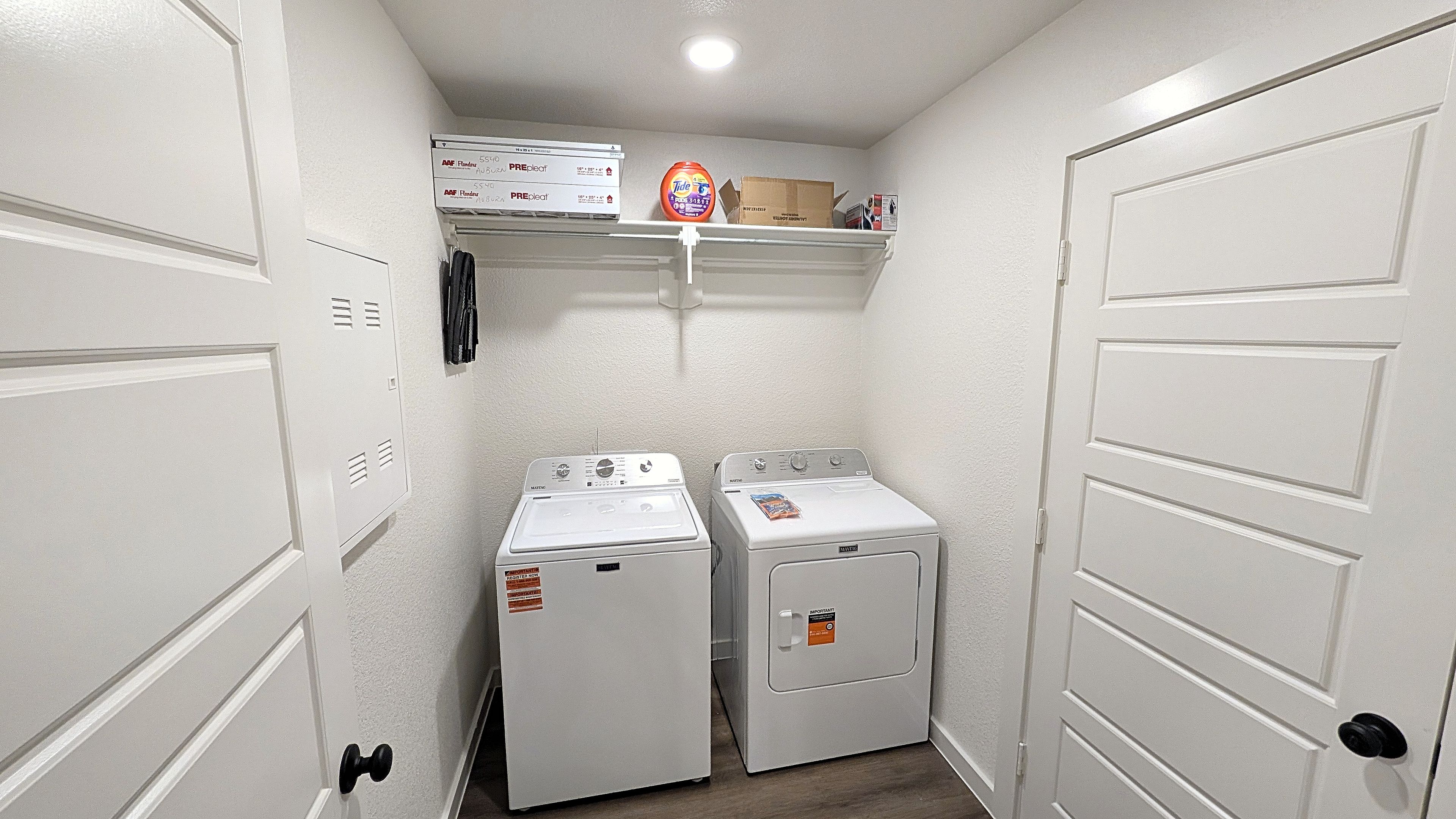 A laundry room with two front-loading washers and dryers, a shelf with laundry supplies, and a white door on the right.