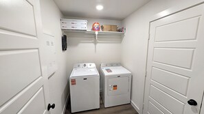 A laundry room with two front-loading washers and dryers, a shelf with laundry supplies, and a white door on the right. - Modern 3BR Auburn St. Retreat (Lubbock)