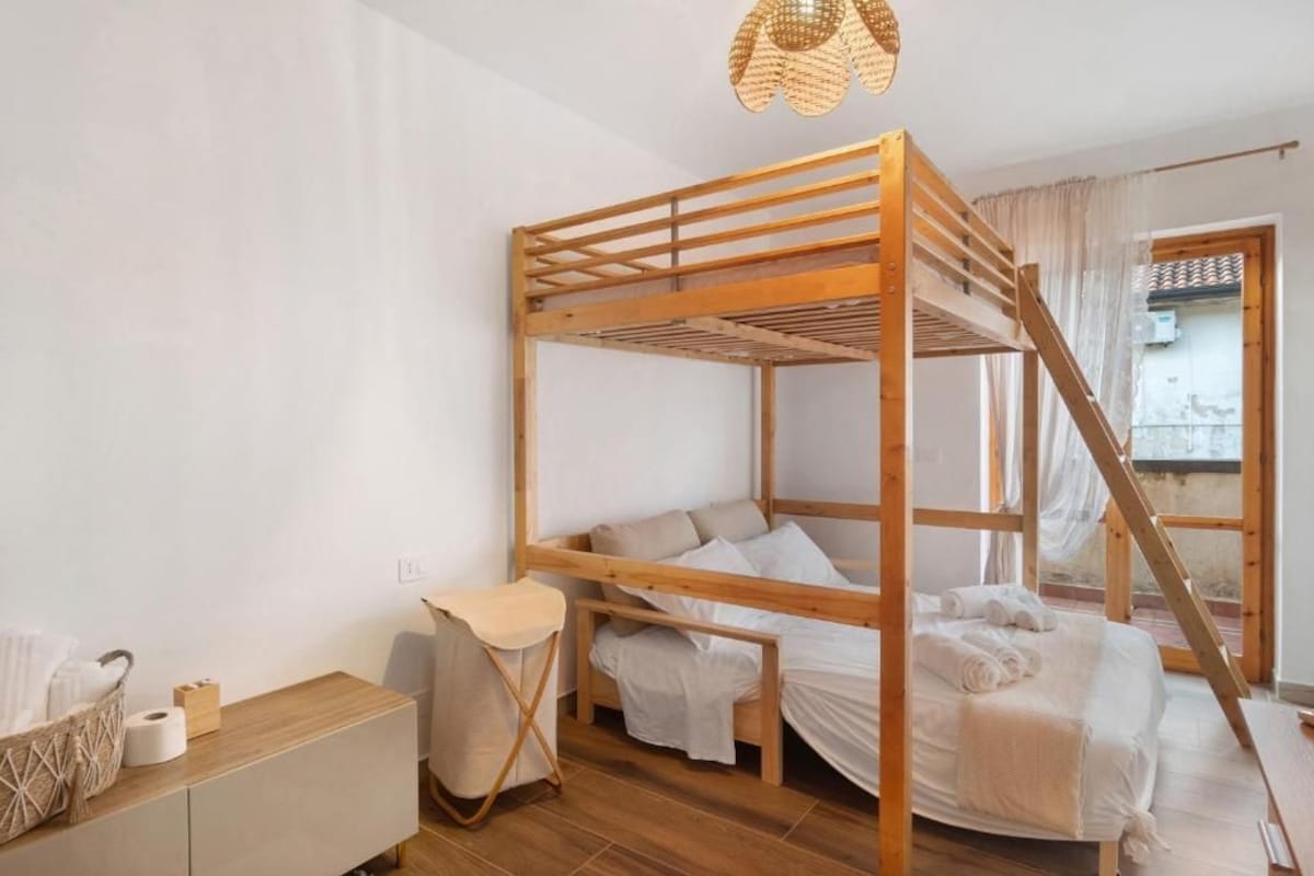 A bunk bed room with a wooden frame, a small bedside table, a chair, and a window with curtains.