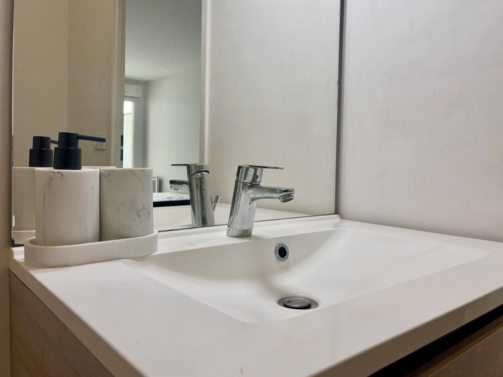 Un lavabo blanc dans la salle de bain, avec un robinet chromé, un miroir au-dessus et un petit récipient contenant des articles de toilette, placé à gauche.