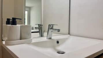Un lavabo blanc dans la salle de bain, avec un robinet chromé, un miroir au-dessus et un petit récipient contenant des articles de toilette, placé à gauche.