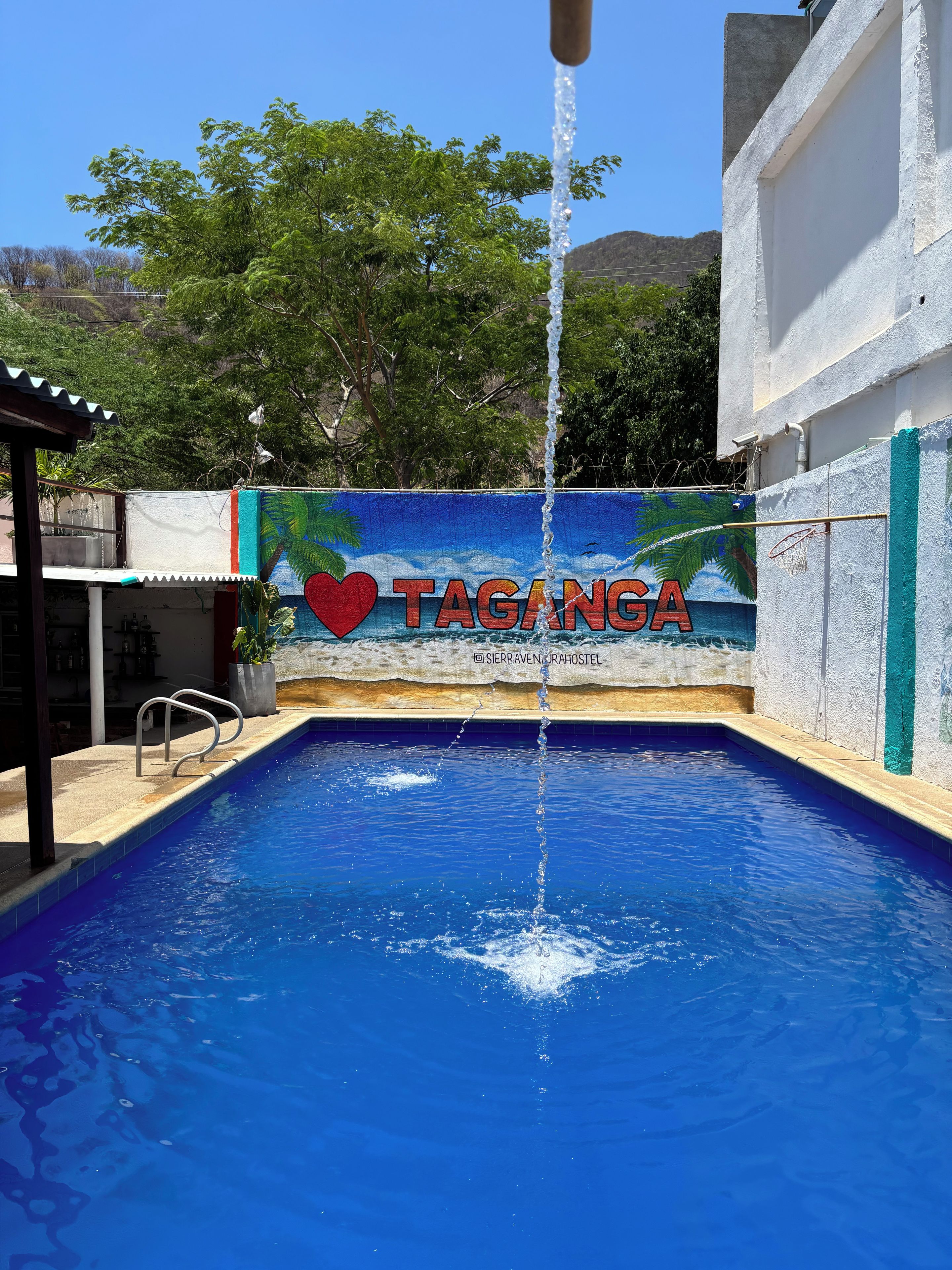 A swimming pool with a water fountain, a mural with the word 'TAGANGA' and a heart symbol, and a view of trees and hills in the background.