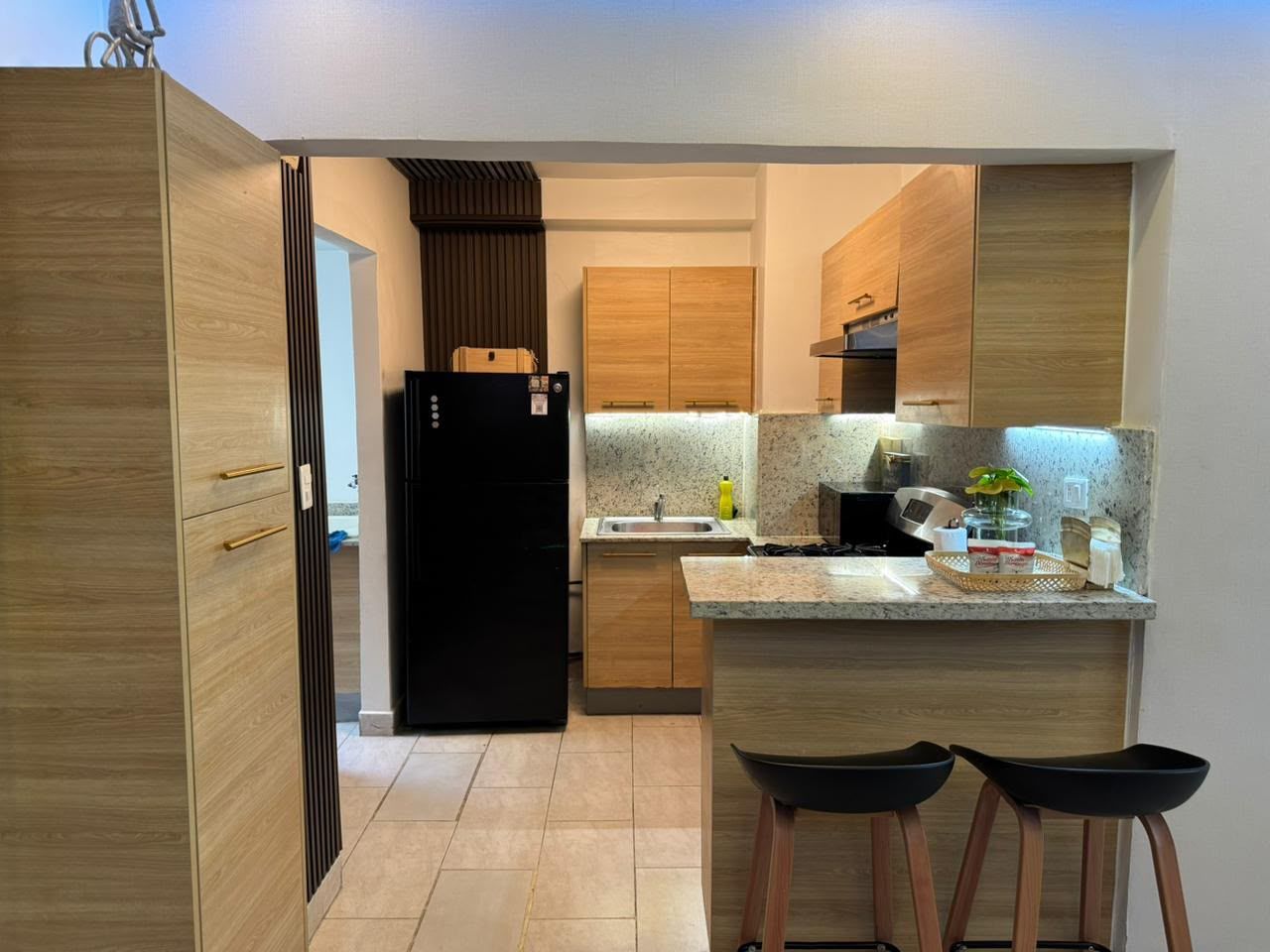 Une cuisine moderne avec des meubles en bois, un réfrigérateur noir, un plan de travail en granit et deux tabourets de bar noirs.
