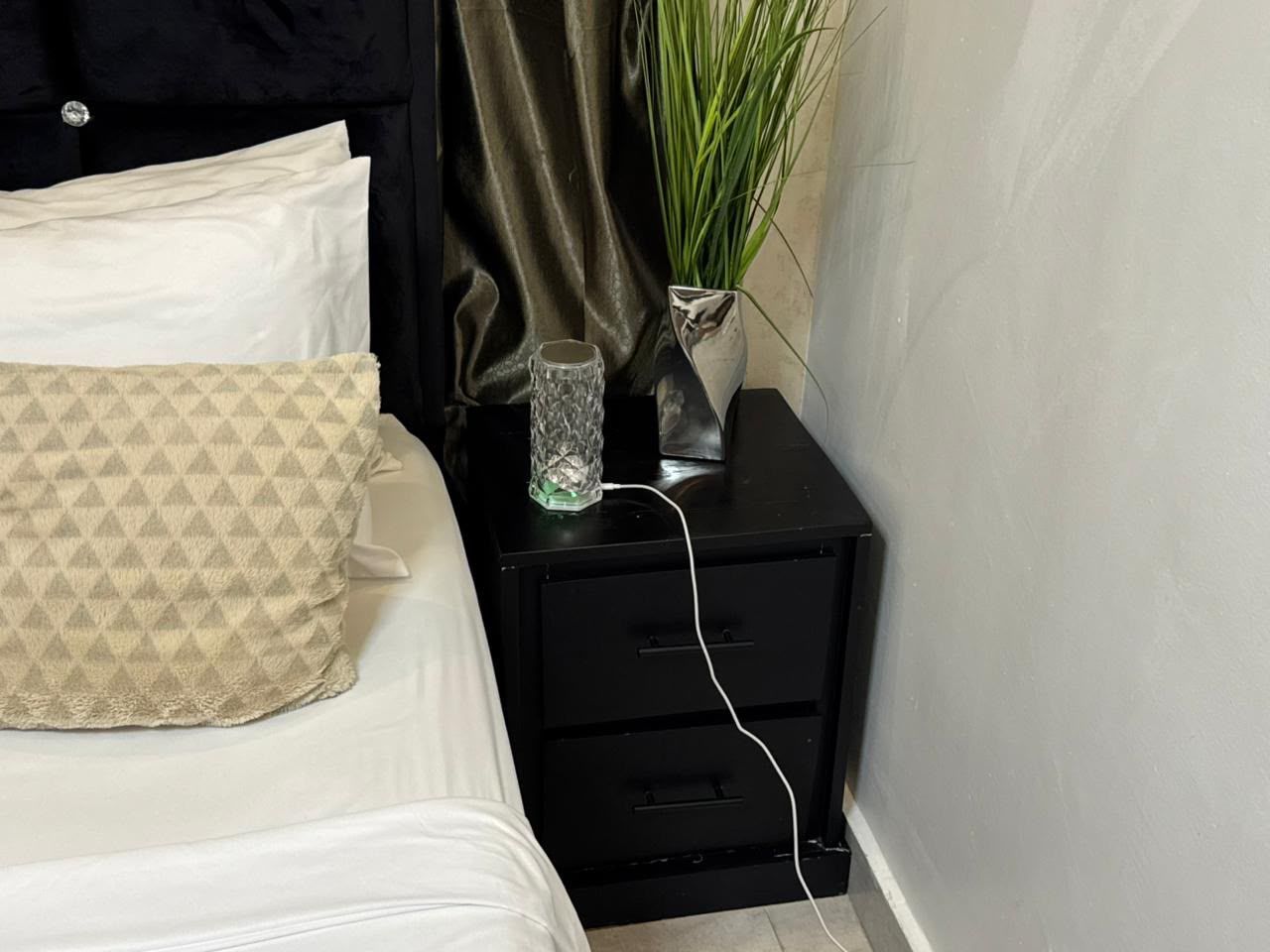 Un lit avec un oreiller à motifs, une table de chevet avec un verre et une plante, et une commode noire avec des tiroirs.