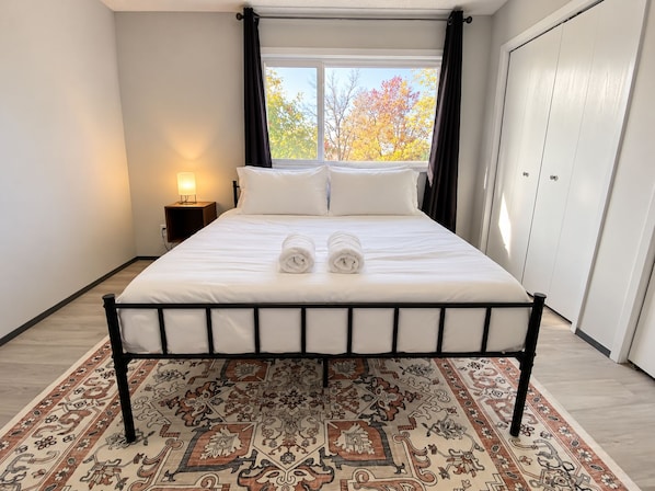 Una cama bien hecha con dos toallas enrolladas, una lámpara de noche y una alfombra estampada en una habitación con ventana y armario.