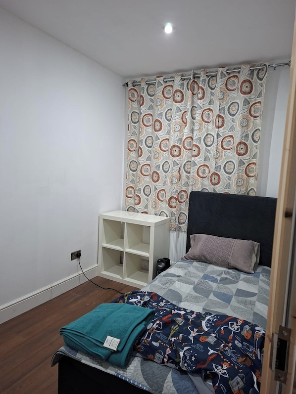 A bedroom with a bed, a bedside table, a camera, and a curtain with a geometric pattern. - House Sleeps 5 With Wifi (Ilford)