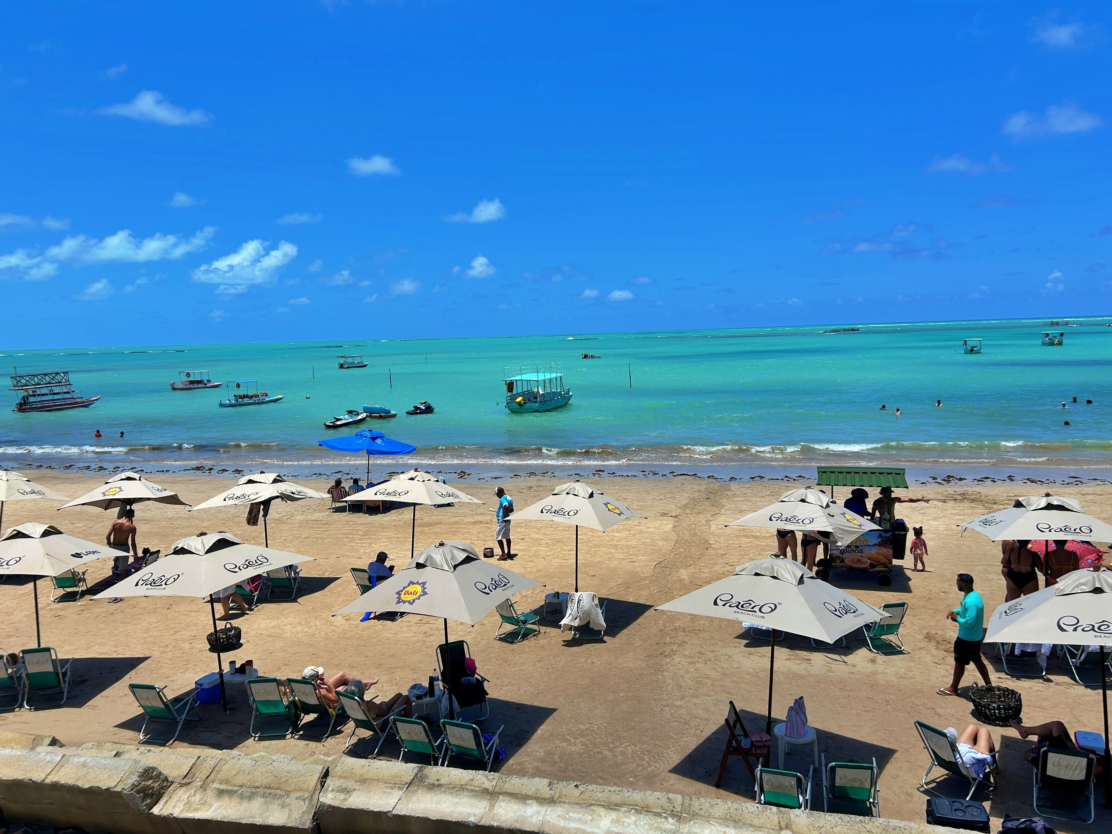 A beach with multiple umbrellas and lounge chairs, a clear blue sky, and boats in the water.